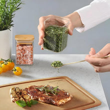 Load image into Gallery viewer, Person seasoning a dish with herbs from a glass container on a kitchen counter.