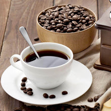 Load image into Gallery viewer, White cup of coffee with a spoon, surrounded by coffee beans and a wooden bowl on a wooden surface.