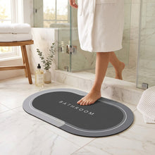 Load image into Gallery viewer, Grey oval bath mat printed BATHROOM, with a foot stepping on it