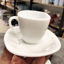 Load image into Gallery viewer, White coffee cup and saucer held in a hand with a blurred store background