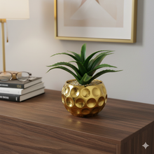 تحميل الصورة في عارض المعرض، Gold decorative pot with a plant on a wooden surface, with books and glasses in the background.