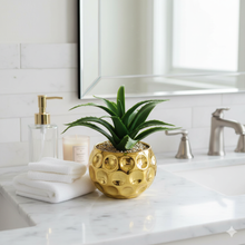 تحميل الصورة في عارض المعرض، Gold pot with a plant on a bathroom counter, next to towels and a candle.