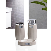 Load image into Gallery viewer, A three-piece bathroom vanity set including a small dish, a tooth holder, and a soap/lotion dispenser, shown in a ribbed beige finish with silver metal tops. The background is a split-screen lifestyle image showing a gray tiled wall and a green plant.