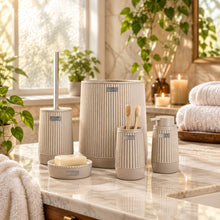Load image into Gallery viewer, A coordinated bathroom accessory set in a light warm gray ribbed ceramic finish, including a toilet brush, holder, cup, soap dish, and bottle, shown in front of a softly blurred bathroom interior.