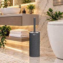 Load image into Gallery viewer, A dark gray toilet brush with a matching handle standing upright in a cylindrical holder, shown in front of a blurred bathroom background with a sink, mirror, and toilet.