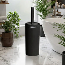 Load image into Gallery viewer, A black toilet brush in a matching cylindrical holder, shown in front of a bathroom scene with a sink, mirror, and toilet.