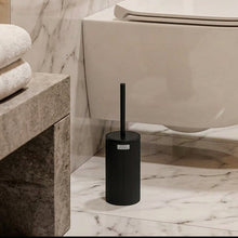 Load image into Gallery viewer, A black toilet brush with a matching cylindrical holder, shown in front of a bathroom scene featuring a sink, mirror, and toilet.