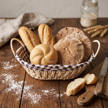 تحميل الصورة في عارض المعرض، A woven bread basket with a brown-and-white gingham liner, filled with assorted breads including baguettes, round rustic loaves, and a round loaf, set on a wooden table with flour, bread slices, a knife, and a cloth napkin.