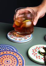 Load image into Gallery viewer, Hand holding a glass of iced drink on a decorative coaster with a dark background