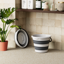 Load image into Gallery viewer, Collapsible gray and white bucket on a tiled floor with a plant and shelves in the background.