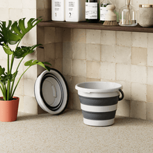 Load image into Gallery viewer, Collapsible gray and white bucket on a tiled floor with a plant and shelves in the background.