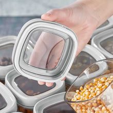 تحميل الصورة في عارض المعرض، A hand holds a square clear plastic spice jar lid with a white rim, shown over a row of matching jars containing dried goods and popcorn kernels.