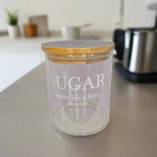 Load image into Gallery viewer, Clear glass jar with wooden lid labeled 'SUGAR makes life a little sweeter' on a kitchen counter.