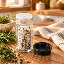 تحميل الصورة في عارض المعرض، A clear glass shaker bottle with a silver metal lid and a separate black plastic cap, placed on a wooden tabletop.