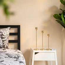 Load image into Gallery viewer, Three slim gold metal candlesticks of varying heights displayed on a white bedside table next to a bed with a black and white botanical patterned headboard and pillow.