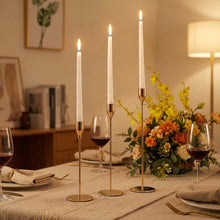 Load image into Gallery viewer, Three slim gold candlestick holders with long white twisted taper candles are arranged on a light blue patterned runner against a white wall. Decorative props include a small amber glass bottle labeled Vissy on the left and yellow flowers on the right.