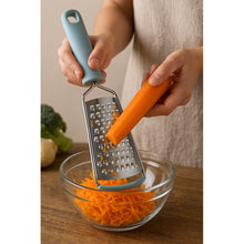 Load image into Gallery viewer, Person using a handheld grater to shred a carrot into a glass bowl on a wooden surface.