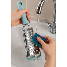 Load image into Gallery viewer, Person washing a metal grater with a blue handle under running water.