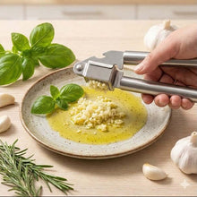 تحميل الصورة في عارض المعرض، Person using a garlic press over a plate of food with herbs and garlic cloves on a wooden surface.