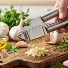 تحميل الصورة في عارض المعرض، Garlic press being used to crush garlic on a wooden cutting board with herbs and oil.