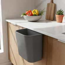 تحميل الصورة في عارض المعرض، A gray under-counter trash bin hanging from a white countertop in a modern kitchen setting with wooden cabinets, a bowl of vegetables, and a small potted plant.