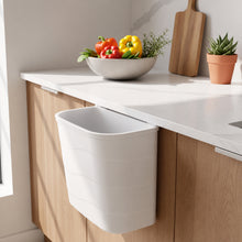 تحميل الصورة في عارض المعرض، A white under-counter hanging trash bin mounted on a light wood kitchen cabinet beneath a white countertop, with a bowl of colorful peppers and a small potted plant nearby.