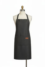 Load image into Gallery viewer, Black apron with a brand logo on a white blanket