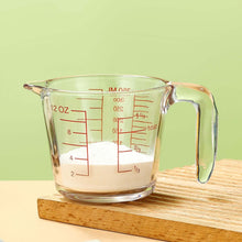 تحميل الصورة في عارض المعرض، Clear glass measuring cup with red measurements on a wooden surface and green background