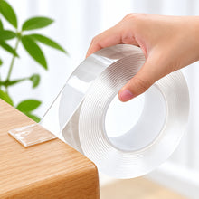 تحميل الصورة في عارض المعرض، A hand peeling back a clear, semi-translucent adhesive strip from a wooden surface, with a roll of similar transparent tape in the background.