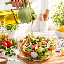تحميل الصورة في عارض المعرض، A hand holds a translucent green squeeze bottle and pours golden oil into a clear glass bowl of salad on a tabletop. The salad contains sliced tomatoes, cucumbers, radishes, greens, and white cheese, with a wooden spoon resting inside the bowl.