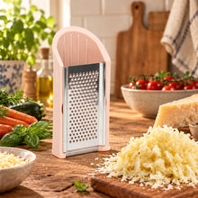 Load image into Gallery viewer, A small handheld cheese grater with a pale pink handle and stainless steel blade, shown on a kitchen counter with garlic bulbs and shredded cheese nearby.