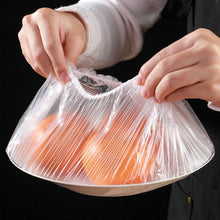 Load image into Gallery viewer, Hands using plastic food storage cover over a bowl of oranges, demonstrating its reusable design for food freshness.