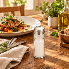 تحميل الصورة في عارض المعرض، Two glass salt and pepper shakers on a white background with a dish of food in the background.
