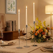 Load image into Gallery viewer, Three tall white taper candles in slim gold metal holders arranged on a dining table, with wine glasses, a floral centerpiece, and warm ambient room lighting in the background.