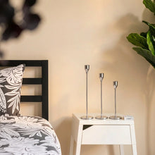 Load image into Gallery viewer, Three slim gold metal candlesticks of varying heights displayed on a white nightstand in a bedroom setting next to a bed with a black headboard and gray and white botanical patterned bedding.