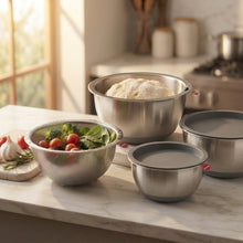 Load image into Gallery viewer, Three stainless steel mixing bowls in different sizes arranged on a kitchen counter, with one bowl containing dough, another filled with mixed greens and tomatoes, and a third bowl beside a dark lid. A cutting board with garlic and herbs sits to the left, and a stove and kitchen cabinets are blurred in the background.