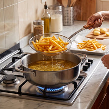 Load image into Gallery viewer, A woman in a kitchen wearing a black apron over a beige long-sleeve shirt, using a slotted spoon to lift french fries out of a pot of oil on a gas stove. More fries are piled on a white plate nearby.