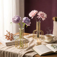 Load image into Gallery viewer, Two gold wire-frame glass vases on a white table, one holding light purple flowers and the other holding pale pink flowers, against a dark burgundy background with decorative books and leaves.