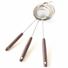 Load image into Gallery viewer, A set of three stainless steel fine mesh round strainers with wooden handles, displayed with the strainers in an upright position and the handles in a curved state.