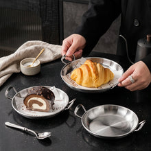 Load image into Gallery viewer, Three small round stainless steel dessert dishes with side handles are arranged on a glossy black surface. One dish holds a croissant, one holds a chocolate cake roll, and one is empty.
