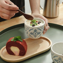 Load image into Gallery viewer, Person holding a bowl of dessert with a red velvet roll cake on a wooden tray.
