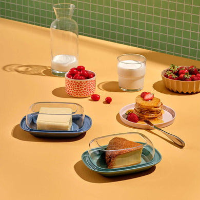A plastic butter dish with an acrylic lid displayed on a table, surrounded by various food items like bread, milk, and strawberries. The dish has a white tray with a transparent cover.