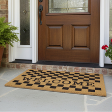 تحميل الصورة في عارض المعرض، Checkered welcome mat in front of a wooden door with plants on either side.
