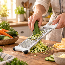 تحميل الصورة في عارض المعرض، A white and stainless steel manual vegetable grater with black rubber tips, resting on a wooden cutting board with sliced potatoes, a cucumber, and a potato in a bright, modern kitchen.
