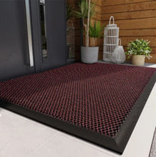 Load image into Gallery viewer, An outdoor doormat with a geometric mesh design in reddish brown and black colors, placed on a tiled floor with a wooden wall in the background.
