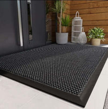 Load image into Gallery viewer, Black textured mat on a white floor with a wooden wall and plants in the background
