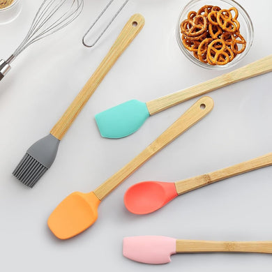 Set of colorful silicone spatulas on a light gray surface with kitchen tools and snacks in the background.