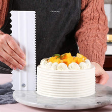 Load image into Gallery viewer, Person decorating a cake with a flexible icing scraper on a marble surface in a cozy kitchen setting.
