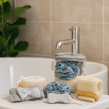 Load image into Gallery viewer, Bathroom setting with various bath products including loofahs and a scrubber on a sink.
