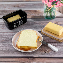Load image into Gallery viewer, Black porcelain butter dish with bamboo lid, spreader, and butter on a plate with flowers in background.
